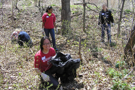 A smiling student volunteer is kneeling with a black trash bag in the foreground while other volunteers search for garlic mustard in the background.