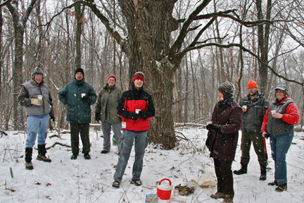 Seven volunteers bundled up in winter clothes stand under a large oak tree holding cups of hot chocolate while it snows.