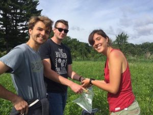 Three college students standing together. All are looking t the camera. One is holding a plastic bag and another is putting a soil sample in it.